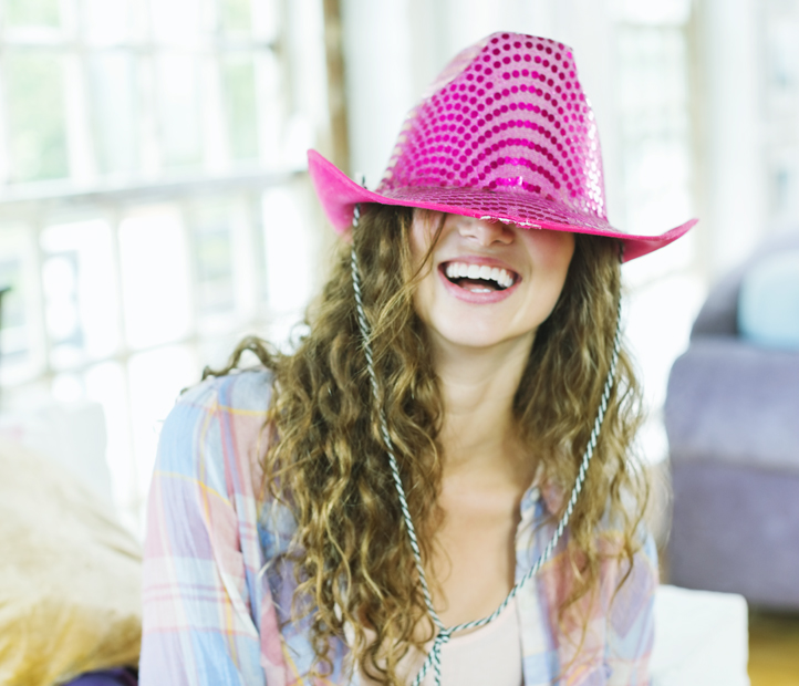 En smilende person med krøllete hår og en glitrende rosa cowboyhatt stråler av glede i et lyst og koselig rom. Bildet uttrykker feiring, energi og optimisme.