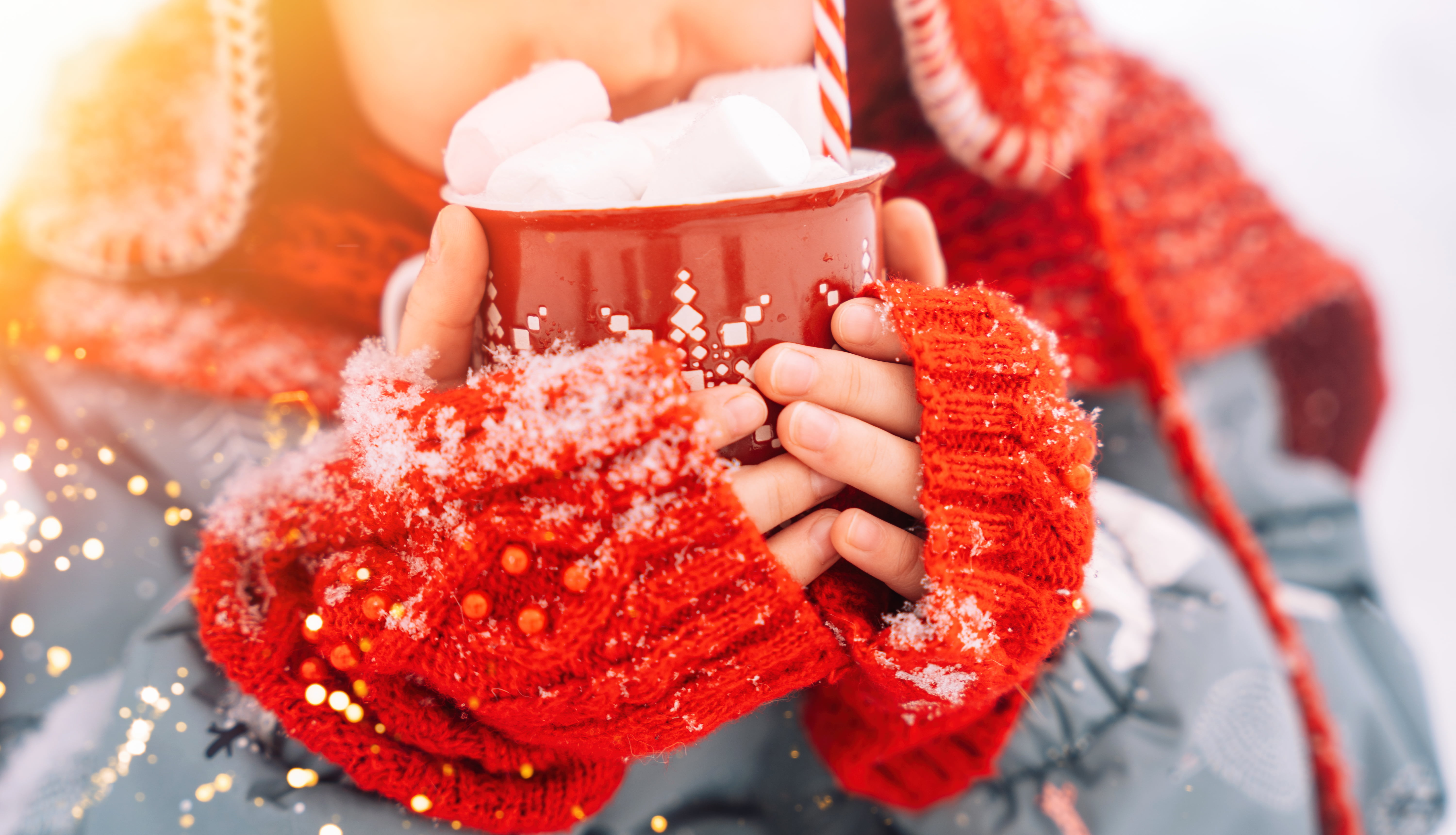 Barn med røde votter som holder en kopp varm kakao med marshmallows ute i snøen.