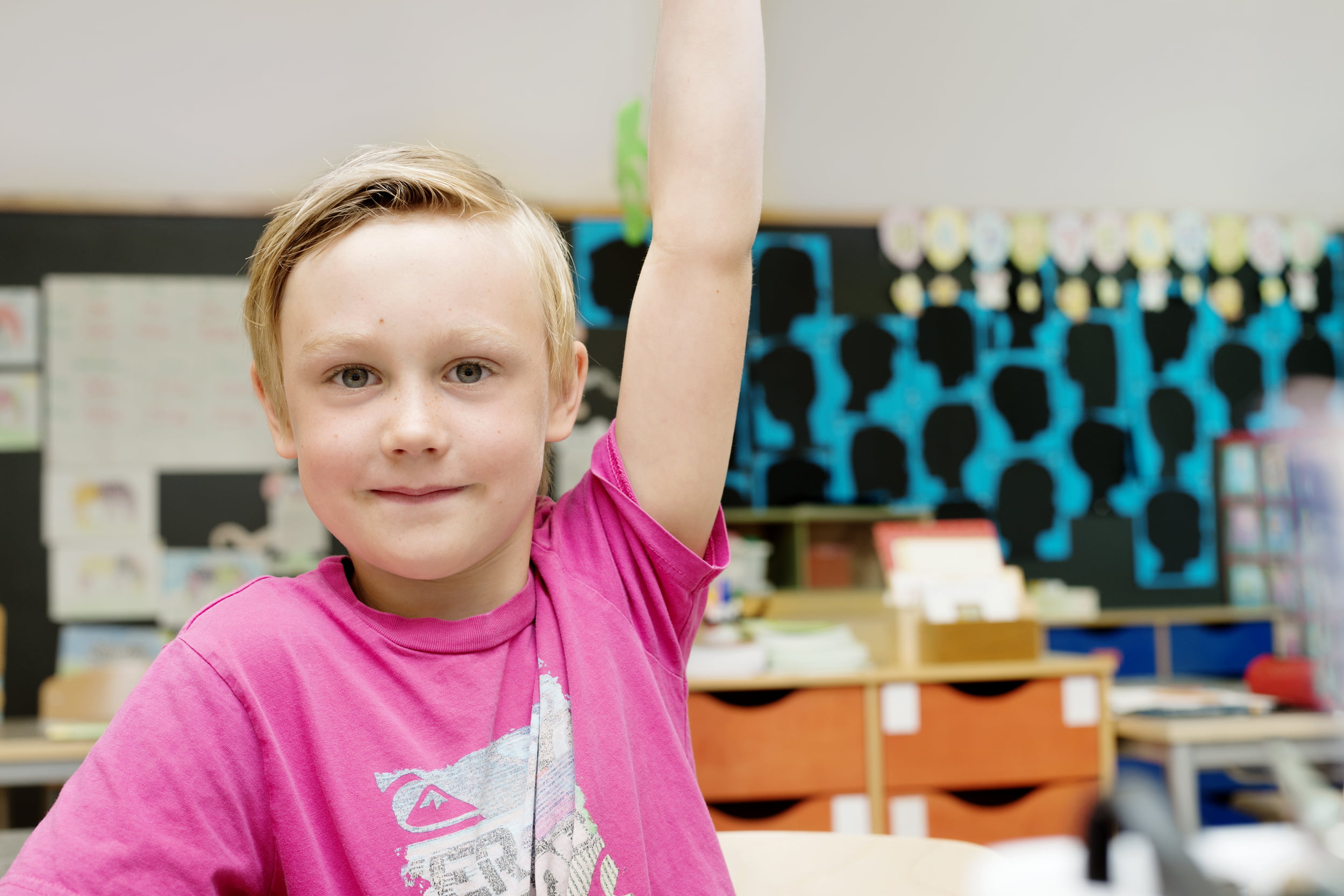 Skolegutt i rosa t-skjorte som rekker opp hånden i et klasserom